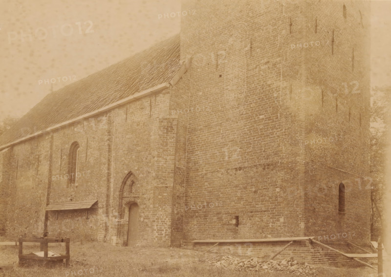 Northwest side of the Reformed Church in Westerwijtwerd, photograph, annotation: 'Westerwijtwerd, municipality of Middelstum', annotation: 'Church in Westerwijtwerd, NW view July 1897, Westerwijtwerd, 1897, photographer, Attributed to, paper, pasteboard, albumen print, height 161 mm x width 218 mm, church (exterior), A large brick building with a steep tiled roof and a tall tower section fills the frame, showing textured masonry, narrow arched windows, and vertical metal rods attached to the walls; a small doorway set into an arched recess and a covered wooden lean-to are visible along one wall, and in front of the building there is a wooden crate-like structure on grass, scattered stones, and a low wooden frame partially covering a shallow brick-lined pit.
