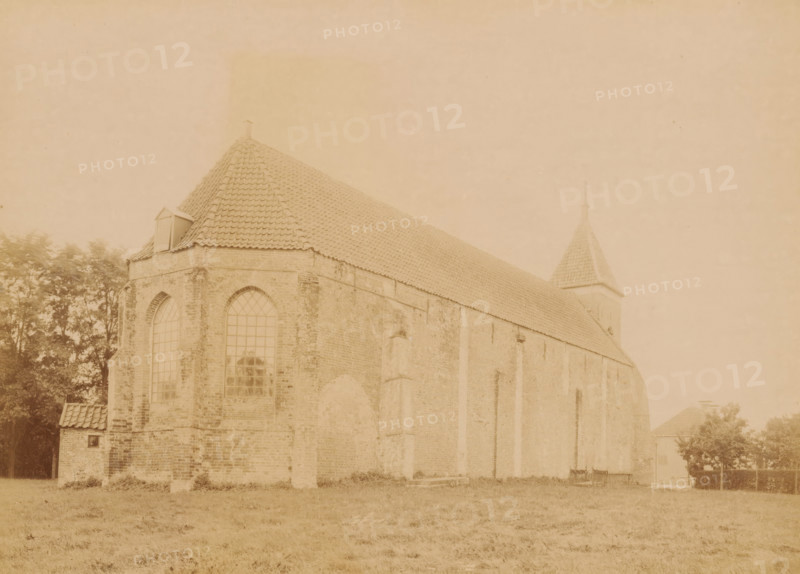 North side of the Church of Vlagtwedde, photograph, annotation: 'Vlagtwedde, north side NH church, Sept 1893', annotation: 'Vlagtwedde', Vlagtwedde, 1893, photographer, Attributed to, paper, pasteboard, albumen print, height 164 mm x width 228 mm, church (exterior), Church of Vlagtwedde, A sepia-toned photograph shows the side and partial front of a large brick building with a steep tiled roof and a small dormer near the roof ridge. Tall arched windows with visible panes are set into the brick wall, and vertical buttresses run along the building's side. A square tower with a pointed tiled roof and a finial rises near the rear. Trees are visible behind and beside the building, and a grassy foreground stretches across the bottom of the image.