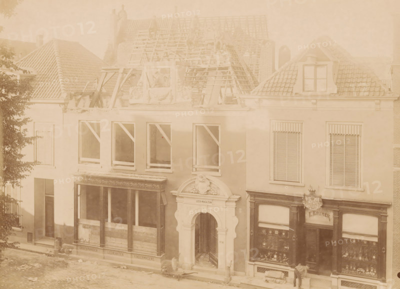 View of Kleine Poot 9 in Deventer, photograph, annotation: 'Deventer, Huis Kleine Poot 9, demolished July 1901', Deventer, c. 1901, photographer, Attributed to, paper, pasteboard, albumen print, albumendruk, height 165 mm x width 229 mm, urban housing, street (city(-scape) with figures, staffage), façade (of house or building), A faded sepia view of a row of adjacent buildings with tiled roofs, dormer windows, and scaffolding on one roof where wooden beams and ladders are visible and a few indistinct figures sit or stand; the façades show multiple window openings with some frames boarded or supported by diagonal braces, decorative stone and plaster ornamentation above a central arched entrance, storefront windows with display cases and decorative moldings, a person standing near one shop entrance, and a tree partially obscuring the left edge of the scene.
