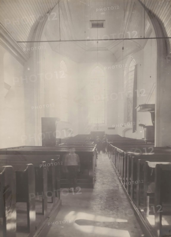 Interior of the Reformed Church in Hellendoorn, photograph, annotation: 'Hellendoorn, Dutch Reformed Church, Sept 1909', Hellendoorn, 1909, photographer, Attributed to, paper, pasteboard, gelatin silver print, height 222 mm x width 163 mm, interior of church, A high, vaulted interior space with wooden pews arranged in rows on either side of a central aisle, tall arched windows along the far wall admitting diffuse light, a curved wooden ceiling with visible paneling and suspended fixtures, a raised pulpit or lectern projecting from the right wall, narrow walkways between pews, and a group of small, blurred figures standing near the back beneath the large windows.