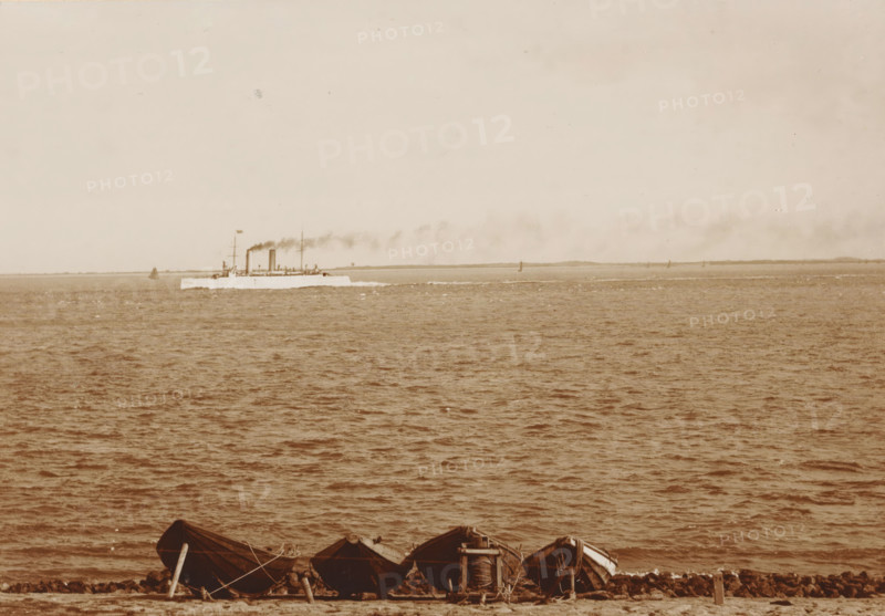 Navy ship Hr. Ms. Zeeland off the coast of Den Helder, 'Zeeland', cabinet photograph, collector's mark: 'Vosmaer', number: '0.50', photographer: Anna Clasina Leijer, Den Helder, 1890 - 1910, Anna Clasina Leijer, photographer, paper, pasteboard, baryta paper, daylight collodion silver print, height 105 mm x width 165 mm, navy, steamship, motorship, sea (seascape), North Sea, A wide view of a calm sea with rippling water filling most of the frame and a pale sky above, a long, low ship steaming along the horizon leaving a trail of dark smoke, several small sail or marker shapes visible near the distant shoreline, and a row of small wooden boats pulled up on a rocky shore in the foreground with ropes and planks nearby.