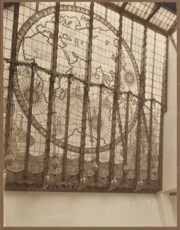 Stained glass window with continents and sailing ships, photograph, check stamp: 'BERN.P.EILERS, AMSTER, DAM', photographer: Bernard Eilers, 1888 - 1951, paper, daylight collodion silver print, height 220 mm x width 170 mm, work of monumental art, maps of the continents and the polar regions, A large decorative window fills the upper portion of the image, composed of many rectangular panes and divided by vertical dark beams; the window contains an intricate circular map-like design with ornate compass roses, stylized coastlines, and sailing ships, rendered in fine linework across the panes. Decorative elements hang from the top of the frame, including patterned bands and suspended geometric lantern shapes, with sculpted figures positioned along the lower edge of the window framework. The lower portion of the photograph shows a plain light-colored wall beneath the window and portions of adjacent architectural surfaces framing the scene. The image has a monochromatic, sepia-toned appeara...