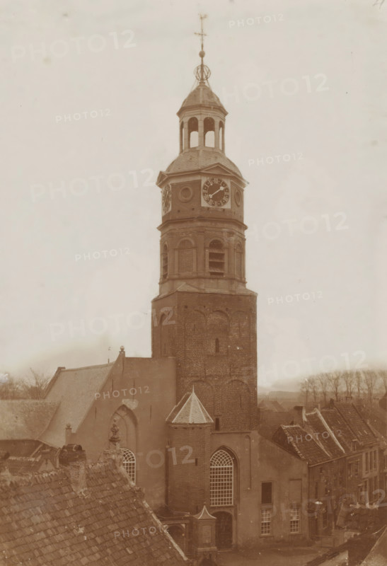 Tower of the St. Lambertus Church in Buren, photograph, annotation: 'NH Kerk, Buren', annotation: 'Tower of the Dutch Reformed congregation in Buren (St. Lambertus)', Buren, 1890 - 1920, photographer, paper, pasteboard, daylight collodion silver print, height 424 mm x width 295 mm, parts of church exterior and annexes: tower, A tall brick church tower rises above surrounding rooftops, topped with a multi-tiered belfry and a domed roof with open arched openings; a clock face is visible on the upper section of the tower, and circular decorative elements flank it; the tower shows arched brickwork and rectangular louvered openings below the clock, and a small conical turret projects from the side of the lower tower above a large arched window with a grid of panes; steep tiled roofs of adjacent buildings and a ridge of rooftops frame the foreground, and rows of bare trees and distant low buildings are visible along the horizon beneath a pale cloudy sky.