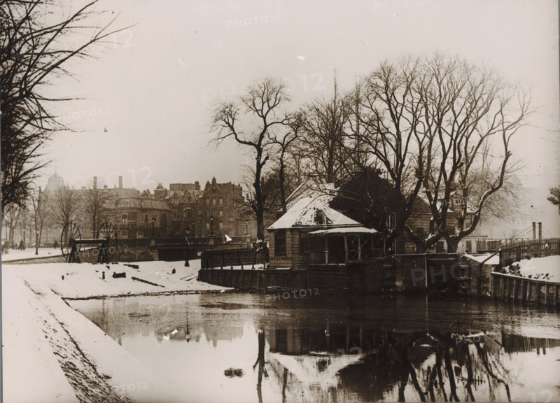 View of a small house on the Stadhouderskade in Amsterdam, photograph, annotation: 'Rijksprentenkabinet, received on loan from mus. Amstelkring, April 1952', annotation: 'Received 10 Nov 1948, glass broken', annotation: 'Small house Stadhouderskade near Rijksmuseum', annotation: '...', Amsterdam, 1900 - 1930, photographer, paper, gelatin silver print, developed gelatin silver print, height 172 mm x width 236 mm, urban housing, street, A snowy riverside scene showing a small wooden building with a pitched roof partially covered in snow, positioned at the water’s edge and surrounded by fencing and wooden posts, with bare trees rising behind and around it. The river reflects the building and trees, and a snow-covered bank slopes down to the water in the foreground. Across the river a bridge or embankment is visible with large spoked wheels mounted on stands nearby, and behind that a row of multi-story stone buildings with chimneys and architectural detailing extends along the skyline. Lea...