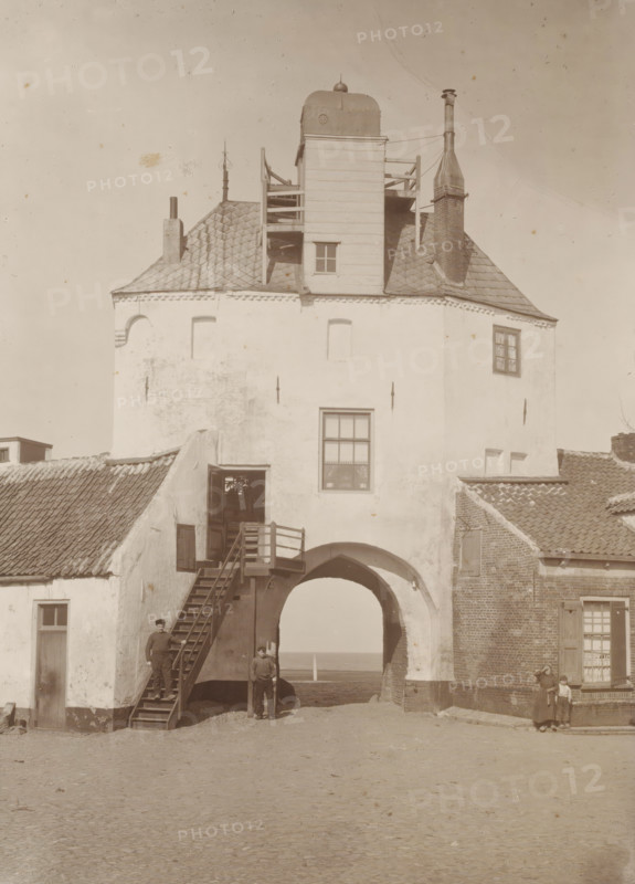 View of the Vischpoort in Harderwijk, photograph, annotation: '1905 Harderwijk, From the Estate of J. Veth, 1972', inscription: 'H. Bickhoff, Amsterdam', photographer: Henri Bickhoff, Harderwijk, 1905, paper, pasteboard, daylight collodion silver print, height 360 mm x width 299 mm, city-gate (city(-scape) with figures, staffage), Vispoort, A tall white plastered building with a tiled roof and chimney features a large arched passage at its base, an external wooden staircase leading to an upper doorway, a square window above the arch, and a wooden tower-like structure rising from the roof with railings and a small window; three men stand near the arch and staircase while a woman and a small child stand near the adjacent brick building, and the ground in front is a cobbled open area.