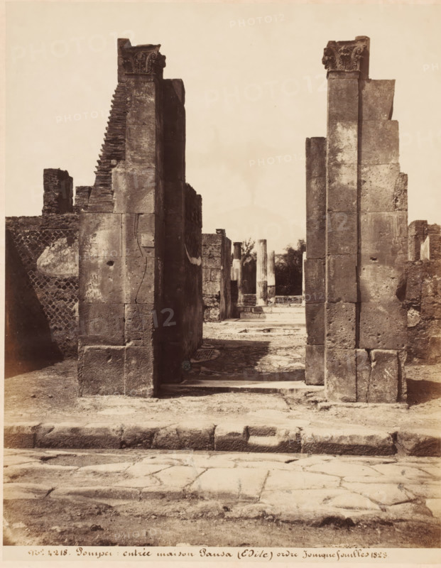 Entrée maison Pansa (Edile) ordre ionique fouillés 1823, Remains of the Casa di Pansa in Pompeii, Italy, Pompei, photograph, collector's mark: 'VOSMAER', number: 'nro. 4218.', photographer: attributed to Giorgio Sommer, Pompeii, 1857 - 1875, Giorgio Sommer, photographer, Attributed to, pasteboard, paper, albumen print, height 306 mm x width 404 mm, urban housing, ruin of a building, architecture, landscape with ruins, Casa di Pansa, A sepia-toned photograph showing a ruined stone structure with two tall, rectangular pillars in the foreground framing a view into an interior space with additional columns and fragmented walls; the pillars have weathered surfaces and ornamental capitals at their tops, and stepped masonry is visible on the left pillar; the ground in front shows a worn stone pavement and a low raised curb, while inside the framed opening there are remnants of fluted columns and scattered architectural debris with vegetation visible beyond; surrounding walls display varying t...