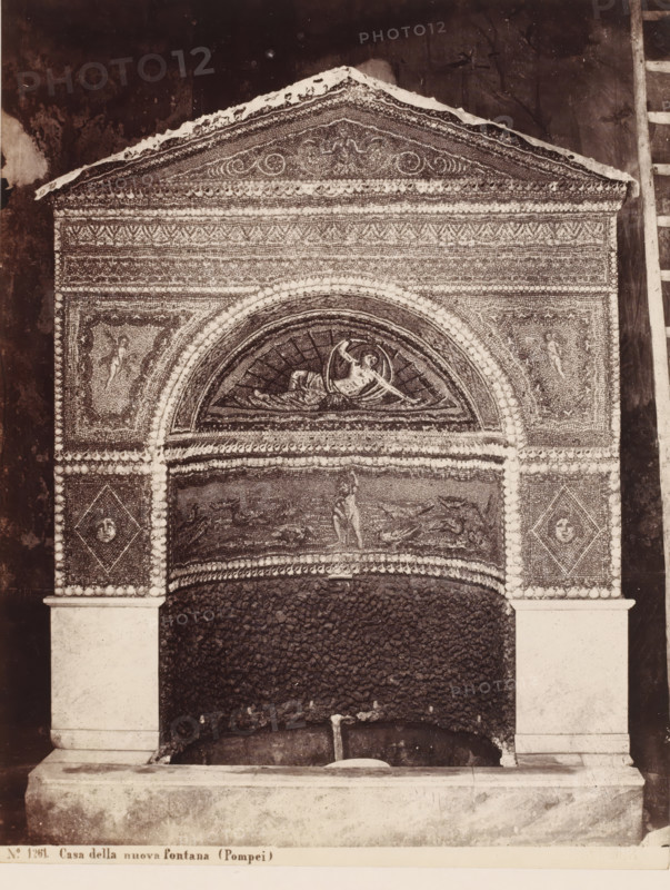 Mosaic in the Casa della nuova Fontana in Pompeii, Italy, Casa della nuova fontana (Pompei), photograph, number: '21', collector's mark: 'RICH SCHOOL FOR ART HANDBOOK, GROUP', number: 'No. 1261.', photographer: Giorgio Sommer, Pompeii, 1857 - 1875, Giorgio Sommer, photographer, pasteboard, paper, albumen print, height 478 mm x width 319 mm, urban housing, ornamental fountain, work of monumental art, Casa della Nuova Fontana, A decorative architectural niche with an ornate triangular pediment sits above an arched recess, the surface covered in dense mosaic patterns and small rounded accents. Inside the arched lunette a reclining figure is depicted within a circular frame, and panels flanking the arch contain smaller human figures. The vertical sides show diamond-shaped mosaic panels with circular motifs, and the lower area beneath the arch is filled with a rough, dark textured surface leading down to a stone basin supported by plain rectangular pilasters. A narrow ledge runs along the b...