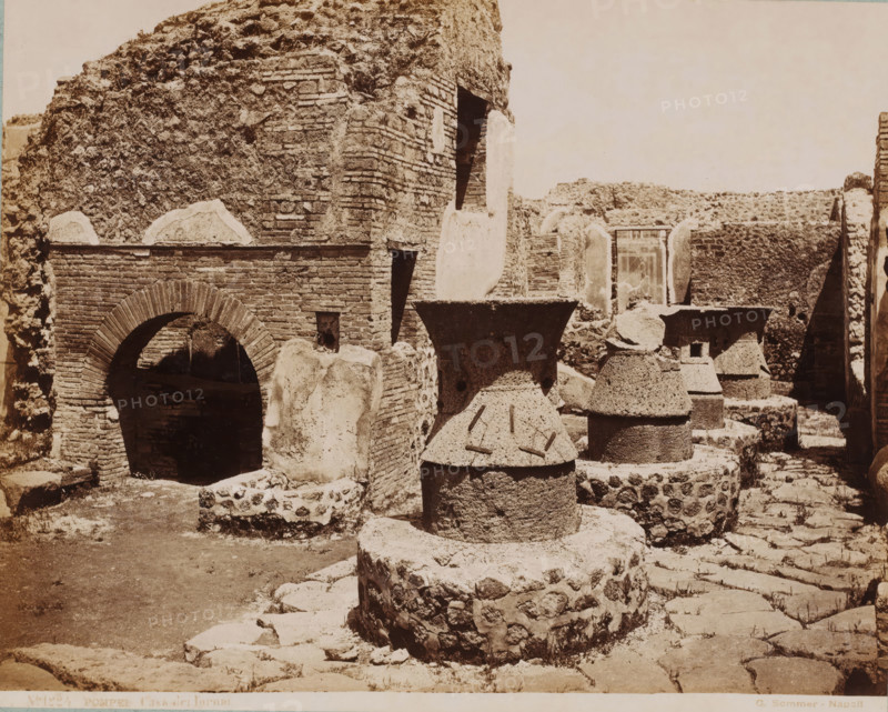 Casa dei Fornai, Remnants of a bakery in Pompeii, Pompei, photograph, number: 'No. 1224.', collector's mark: 'VOSMAER', photographer: Giorgio Sommer, Pompeii, 1863 - 1875, Giorgio Sommer, photographer, pasteboard, paper, albumen print, height 315 mm x width 420 mm, shop, store, ruin of a building, architecture, landscape with ruins, bread, loaf, A sepia-toned photo shows a ruined stone and brick structure with rough textured walls and arched openings. In the foreground there are large cylindrical stone mills with conical upper parts and circular bases, some showing grooves and embedded metal pieces. A prominent brick archway leads into a dark interior space on the left side. Scattered stone blocks and low retaining walls form uneven ground and steps, and additional stone and brick walls with rectangular openings and rounded tops appear in the middle and background. The scene is bathed in soft, even light and contains worn surfaces, rubble, and patches of exposed mortar.