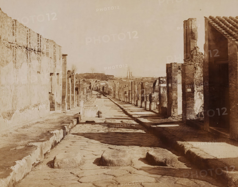 Strada della Abbondanza, View of the Strada della Abbondanza in Pompeii, Italy, Pompei, photograph, number: '1262', collector's mark: 'VOSMAER', photographer: Giorgio Sommer, Pompeii, 1857 - 1875, Giorgio Sommer, photographer, pasteboard, paper, albumen print, height 315 mm x width 420 mm, street, ruin of a building, architecture, landscape with ruins, Strada dell' Abbondanze, A long ancient street lined with ruined stone and brick walls and fragmented columns stretches into the distance under a pale sky, with a raised paved sidewalk on one side and a narrow curb on the other, uneven cobblestones covering the road and several rounded stone stepping blocks in the foreground, remnants of tiled roofs and a partially collapsed doorway visible on the right, and low earthen mounds and sparse bare trees on the horizon.