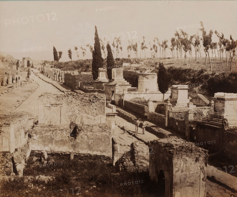 View of the Strada delle Tombe in Pompeii, Italy, Strada delle Tombe, Pompei, photograph, collector's mark: 'VOSMAER', number: '1278', photographer: Giorgio Sommer, Pompeii, 1857 - 1875, Giorgio Sommer, photographer, pasteboard, paper, albumen print, height 315 mm x width 422 mm, street, ruin of a building, architecture, landscape with ruins, gravestone, tombstone, cemetery, park-like burying ground, Via delle Tombe (Pompeï), A wide view of an ancient ruined street flanked by crumbling stone walls and partially standing structures, with paved pathway running through the center, scattered rectangular tomb-like monuments and raised stone platforms along one side, tall narrow cypress trees and a line of thinner trees visible on the ridge beyond the ruins, grassy patches and rubble filling open areas, two figures walking on the paved path, and the scene rendered in a sepia-toned photographic style.