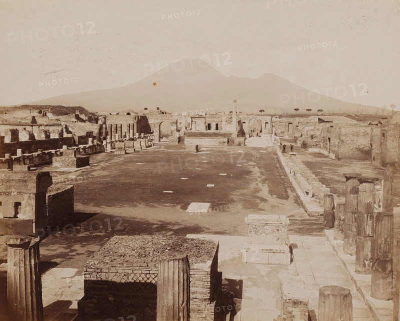 View of the Forum in Pompeii, Pompei, photograph, number: '1202.', collector's mark: 'VOSMAER', photographer: attributed to Giorgio Sommer, Pompeii, 1863 - 1875, Giorgio Sommer, photographer, Attributed to, pasteboard, paper, albumen print, height 314 mm x width 421 mm, In the background right of the square another photographer is at work., portrait, self-portrait of photographer, square, circus, ruin of a building, architecture, landscape with ruins, Piazza del Foro, A wide sepia-toned view of ancient ruins with rows of stone columns, broken walls, and rectangular stone blocks stretching into the distance, an open paved central area with scattered square stones, a raised platform and archways toward the middle ground, two small figures standing near a paved walkway at the far right, low vegetation and a line of trees visible beyond the ruins, and a large gently sloping mountain forming the background under a pale sky.