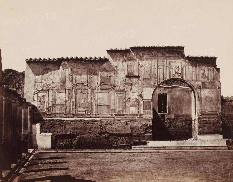 Bagni nuovi (Pompeii), Ruins of a bathhouse in Pompeii, photograph, number: '28', inscription: 'Pompei', number: '29', collector's mark: 'RICH SCHOOL FOR ARTS HANDBOOK, GROUP', number: 'No. 1258.', photographer: attributed to Giorgio Sommer, Pompeii, 1863 - 1875, Giorgio Sommer, photographer, Attributed to, pasteboard, paper, albumen print, height 320 mm x width 478 mm, ruin of a building, architecture, landscape with ruins, public baths, Bagni nuovi, A sunlit facade of an old stone building fills the middle of the image, showing faded decorative panels and reliefs, a large arched opening with a dark interior, and a rectangular window above a worn wall; tiled roof eaves cast shadows along the top edge and small sculpted figures appear near the arch; the lower portion of the wall shows exposed brick and rough texture, with a stepped platform and a shallow paved foreground in front; on the left side a wooden fence and a small plastered structure are visible, and long diagonal shadows fal...