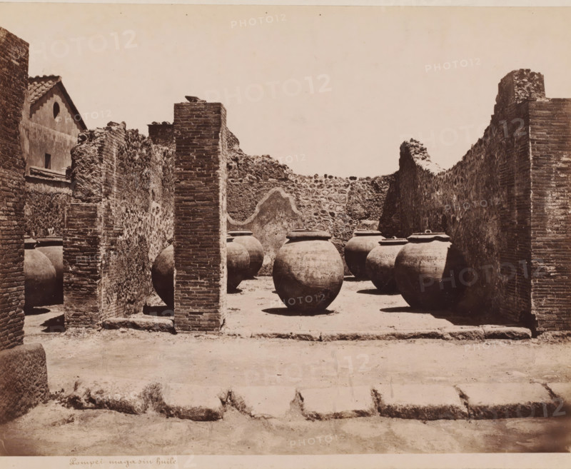 Remains of a shop with oil jars in Pompeii, Pompei magasin huile, photograph, collector's mark: 'VOSMAER', photographer: attributed to Giorgio Sommer, Pompeii, 1863 - 1875, Giorgio Sommer, photographer, Attributed to, pasteboard, paper, albumen print, albumendruk, height 306 mm x width 404 mm, shop, store, ruin of a building, architecture, A sunlit courtyard enclosed by stone and brick walls contains a row of large rounded clay jars with lids resting on the ground, some partially in shadow; a tall brick pillar stands in the foreground between the jars and a rough textured wall, and remnants of masonry and a building facade with a circular opening are visible at the left, while the right side shows a higher brick wall with uneven top edges and a section of worn paving in front.