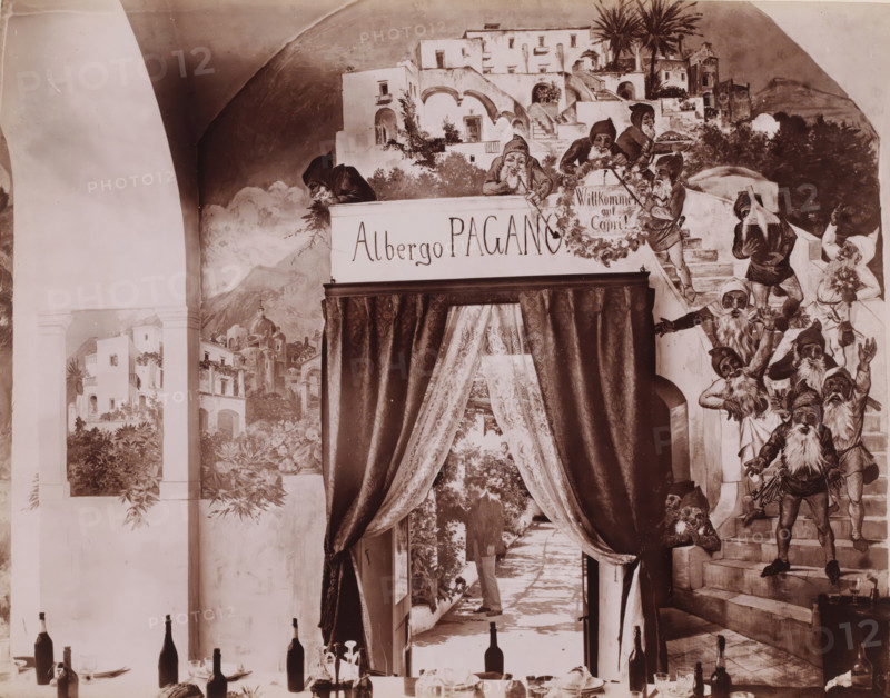 Interior of the dining room of Hotel Pagano on Capri, Italy, Capri, photograph, inscription: 'Capri', number: '6125', photographer: attributed to Giorgio Sommer, 1857 - 1914, Attributed to, pasteboard, paper, albumen print, height 317 mm x width 445 mm, hotel, hostelry, inn, Isola di, A painted wall scene with architectural elements showing arched openings, stairways, terraces, and clustered white buildings surrounded by foliage, palm trees, and distant hills, populated by many small bearded figures wearing pointed hats and tunics, some standing on steps and others leaning over balconies, a heavy dark curtain with patterned drapery framing a passageway that leads to a sunlit courtyard with potted plants and a person partially visible in the distance, a dining setup in the foreground with bottles, glasses, plates, cutlery, and a few decorative items arranged along a table edge.