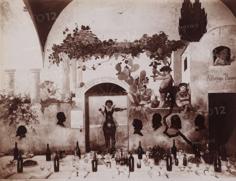 Hotel Pagano, Interior of the dining room of Hotel Pagano in Capri, Italy, Capri, photograph, number: '5170 ?', photographer: attributed to Giorgio Sommer, 1857 - 1914, Attributed to, pasteboard, paper, albumen print, height 318 mm x width 445 mm, hotel, hostelry, inn, Isola di, A painted wall scene shows an archway with climbing vines and leaves overhead, a cactus plant with pads, and sculpted columns. Several cherub-like figures and children are depicted in various poses around the arch and a small building facade, one child holding a platter and another leaning while playing a small instrument. A central painted figure sits astride a donkey visible through an opening beneath the vines. Silhouetted profile heads appear arranged across the lower portion of the wall. In front of the wall a long table is set with plates, folded napkins, glasses, bottles, serving dishes, and floral arrangements, with scattered utensils and a loaf of bread on a platter. The overall palette is muted and se...