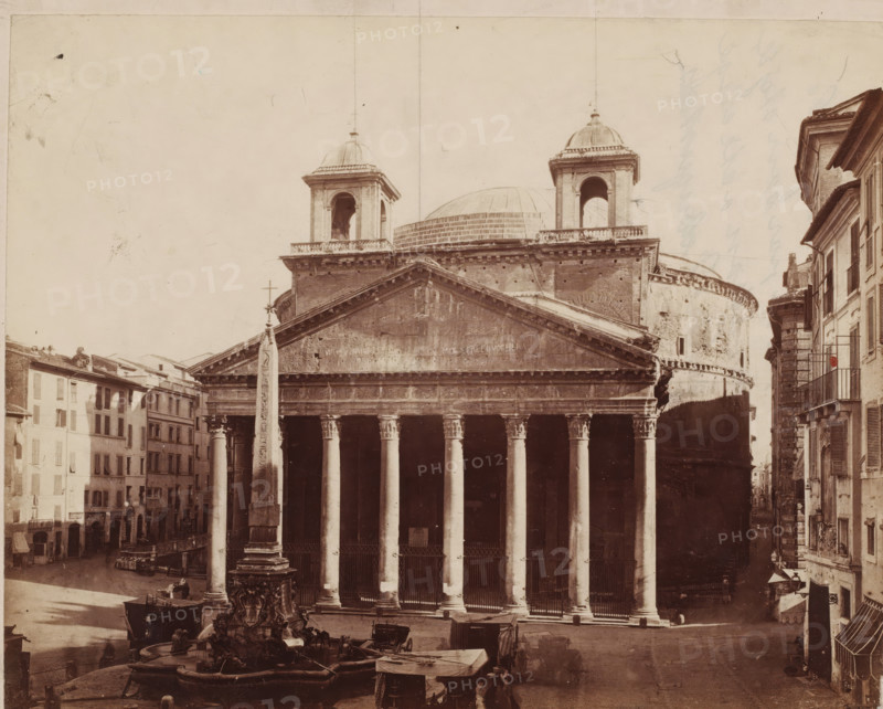 View of the Pantheon and the Macuteo obelisk in Rome, Italy, photograph, inscription: 'Leskast pl. I', inscription: 'Chagrijn, M. Chagrijnig, ...', collector's mark: 'RIJKS SCHOOL FOR ART AND HANDY, GROUP', inscription, photographer: attributed to Giorgio Sommer, Rome, after sculpture by anonymous, 1857 - 1914, Giorgio Sommer, photographer, after sculpture by, Attributed to, pasteboard, paper, albumen print, height 320 mm x width 479 mm, square, circus, church (exterior), obelisk, needle, Pantheon, Piazza della Rotonda, A wide sepia-toned photograph shows a classical temple facade with a triangular pediment and a row of tall fluted columns supporting an entablature, flanked by two square bell towers topped with domes and a large rounded dome visible behind them; in front of the temple stands an obelisk mounted on an ornate fountain base with sculpted figures and water basins, horse-drawn carriages and covered carts are parked nearby, multi-story buildings with shuttered windows line th...