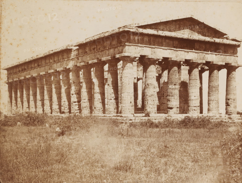 Tempio di Nettuno, Temple of Neptune, Paestum, Italy, Pesto, photograph, number: 'No. 4116 ?', collector's mark: 'VOSMAER', photographer: attributed to Giorgio Sommer, 1857 - 1914, Giorgio Sommer, photographer, Attributed to, pasteboard, paper, albumen print, albumendruk, height 319 mm x width 478 mm, ruin of a building, architecture, temple, shrine, non-Christian religions, Second Temple of Hera, A wide sepia-toned photograph of a large ancient stone temple with a long row of fluted columns supporting an entablature and a pitched roof, the columns showing weathering and irregular surface texture, shadows cast between them, and gaps where inner spaces are visible; the temple sits on a raised platform with a rough foreground of grass and low vegetation extending toward the viewer, and the sky above is pale with scattered small blemishes on the photographic surface.