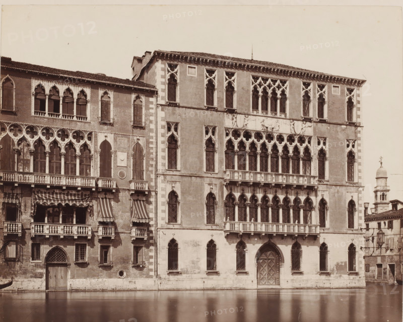 View of the Palazzo Foscari in Venice, Italy, Palazzo Foscari (Venezia), photograph, inscription: 'Venetie, Venetia', collector's mark: 'RICH SCHOOL FOR ART HANDBOOK, GROUP', inscription: 'I', number: 'No. 3532.', number: '41', photographer: attributed to Giorgio Sommer, Venice, 1857 - 1914, Giorgio Sommer, photographer, Attributed to, pasteboard, paper, albumen print, albumendruk, height 319 mm x width 480 mm, hôtel, ie small palace in city, Ca' Foscari, A sepia-toned photograph of an ornate stone building facade facing a canal, showing multiple levels of arched windows and balconies with decorative columns and tracery, a large carved central doorway with a pointed arch, striped fabric awnings over some balcony openings, patterned window grilles, rectangular and arched window openings with stone surrounds, sculpted relief panels and ornate cornices near the roofline, a narrow vertical section with lighter masonry at a corner, and a bell tower with a domed top visible beyond the buildi...