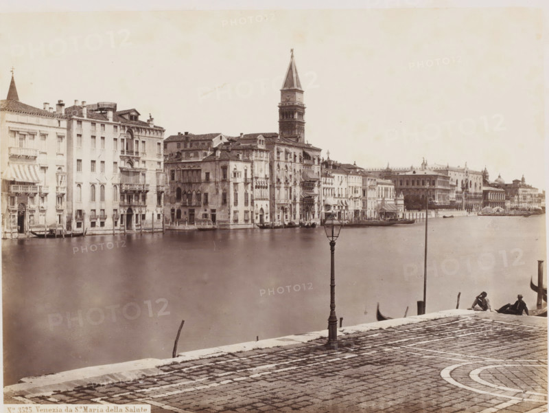 View of the Grand Canal and the Sante Maria della Salute in Venice, Italy, Venezia da Sa Maria della Salute, photograph, address: 'GIORGIO SOMMER, STUDIO MONTE DI DIO 4., MAGAZZINO S. GATERINA 5., NAPOLI', number: 'No. 3525.', photographer: Giorgio Sommer, Venice, 1857 - 1914, pasteboard, paper, albumen print, height 304 mm x width 375 mm, canals, waters (in city), church (exterior), Basilica of Santa Maria della Salute, Grand Canal, A wide waterfront view shows a row of multi-story stone buildings with varied facades and balconies lining a canal, several with awnings and small boats moored at water-level entrances; a tall bell tower rises behind the buildings toward the center of the scene; the foreground features a cobbled promenade with a decorative curved paving pattern, a tall ornate lamppost near the water, and two seated figures at the edge beside a small boat; smooth water occupies the middle ground between the promenade and the buildings.