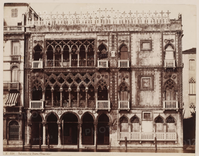 View of Palazzo Cà d'Oro in Venice, Italy, Palazzo Cà Doro. (Venezia), photograph, number: '46', inscription: 'Venetie', collector's mark: 'RICH SCHOOL FOR ART HANDBOOK, GROUP', number: 'No. 3530.', inscription: 'I', photographer: attributed to Giorgio Sommer, Venice, 1857 - 1914, Attributed to, pasteboard, paper, albumen print, height 319 mm x width 480 mm, hôtel, i.e. small palace in city, Ca' d'Oro, A sepia-toned photograph of an ornate stone facade facing water, with a ground-level arcade of rounded arches supported by columns, above which sit rows of Gothic windows with pointed arches and intricate tracery, some windows opening to small balustraded balconies. The upper cornice is topped by a decorative crenellated parapet with sculpted finials, and the facade surface displays patterned stonework, carved medallions, and framed panels with relief details. Flanking sections show vertical twisted columns and narrow windows with shutters, while the lower edge of the building meets a ca...