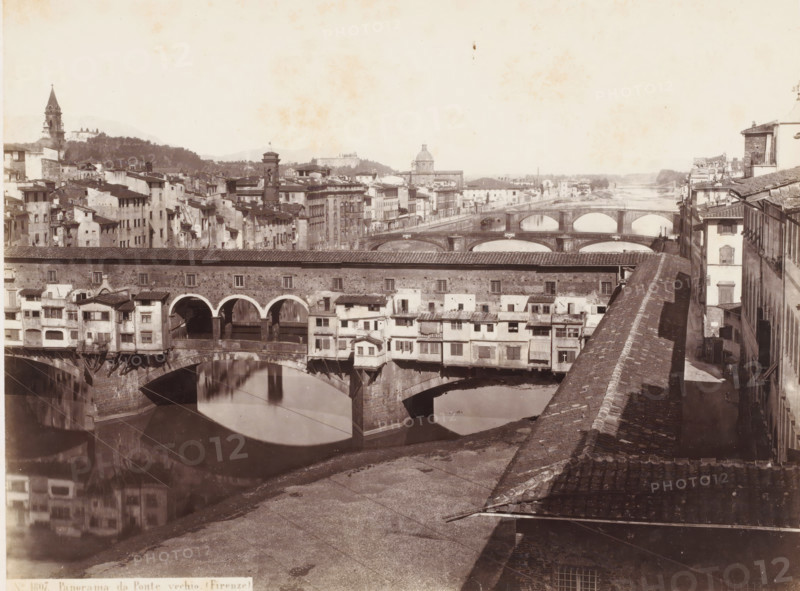 View of the Arno and the Ponte Vecchio in Florence, Italy, Panorama da Ponte vechio. (Firenze), photograph, address: 'DEPOT A FLORENCE, J. BRECKER, VIA MAGGIO 15', address: 'GIORGIO SOMMER, STUDIO MONTE DI DIO 4., MAGAZZINO S. GATERINA 5., NAPOLI', number: 'No. 1807.', photographer: Giorgio Sommer, Florence, 1857 - 1914, Tuscany, pasteboard, paper, albumen print, height 305 mm x width 377 mm, landscape with bridge, viaduct or aqueduct, river, bridge in city with structures, ie shops, houses, eg Ponte Vecchio in Florence, Ponte Vecchio, Arno, A wide river scene with multiple stone bridges crossing the water, the nearest bridge featuring arches and small buildings projecting from its sides, their facades showing windows, shutters, and tiled roofs; the water below reflects the arches and buildings. Along the riverbanks there are rows of closely packed multi-story buildings with varied rooflines and external stairways, and a long roofed structure running parallel to the river in the midgro...