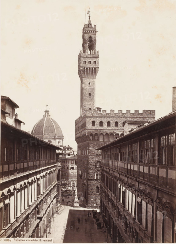 View of the Uffizi Gallery, Palazzo Vecchio and the Cathedral in Florence, Italy, Palazzo Vecchio. (Firenze), photograph, address: 'DEPÔT A FLORENCE, J. BRECKER, VIA MAGGIO 15', number: 'No. 3804.', address: 'GIORGIO SOMMER, STUDIO MONTE DI DIO 4., MAGAZZINO S. CATERINA 5., NAPOLI', photographer: Giorgio Sommer, Florence, 1857 - 1914, Tuscany, pasteboard, paper, albumen print, height 374 mm x width 305 mm, permanent exhibition, museum, church (exterior), townhall, Palazzo Vecchio, Florence Cathedral, Uffizi Gallery, A narrow cobblestone street flanked by two long multi-story buildings with rows of windows and covered walkways leads toward a tall stone tower rising above a fortified palace facade; the tower has a crenellated upper section, arched openings near its top, and a decorative spire. In the distance a large domed building with a ribbed surface is visible behind the tower. A statue stands on a pedestal in the open area at the base of the tower, and small groups of people and hor...