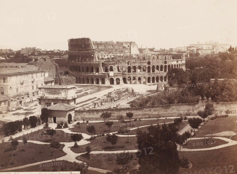Colosseo preso dai giardini farnesi (Roma), View of the Colosseum in Rome, Italy, photograph, number: '7.', address: 'SOMMER & BEHLES, ROMA E NAPOLI', number: 'No. 1024.', inscription: 'Roma', inscription: 'C. 85', photographer: Sommer & Behles, Rome, 1867 - 1874, pasteboard, paper, albumen print, albumendruk, height 320 mm x width 378 mm, ruin of a building, architecture, amphitheatre, Colosseum, A wide sepia-toned view of an ancient oval stone amphitheater with multiple levels of arched openings, showing weathered and partially ruined exterior walls; in front of it a low rectangular stone structure with a flat top and a building with arched windows and a tiled roof; winding pale pathways curve through a grassy garden area dotted with small trees and shrubs across the foreground; dense trees and taller vegetation occupy the area to the right of the amphitheater and extend into the background; scattered low buildings and ruins appear beyond and to the left of the amphitheater under a p...