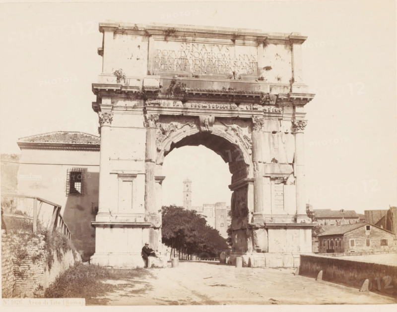 Arco di Tito (Roma), Triumphal Arch of Titus in Rome, Italy, photograph, number: '6', number: 'No. 1026.', inscription: 'C. 85.', inscription: 'Roma', address: 'SOMMER & BEHLES, ROMA E NAPOLI', photographer: Sommer & Behles, Rome, 1867 - 1874, pasteboard, paper, albumen print, albumendruk, height 321 mm x width 380 mm, ruin of a building, architecture, triumphal arch, gate, entrance, Arch of Titus, A large stone archway with ornate carvings and columns dominates the scene, showing weathering and plant growth on its upper surfaces and decorative friezes. A person wearing a hat sits near one of the arch bases, holding a long object and positioned on a small raised platform. Behind the arch a row of trees lines a path leading toward a distant tower visible through the arch opening. To the left of the arch a building with a tiled roof and a barred window sits adjacent to a sloped stone embankment and wooden stairs. To the right of the arch smaller stone buildings and rooftops extend toward...