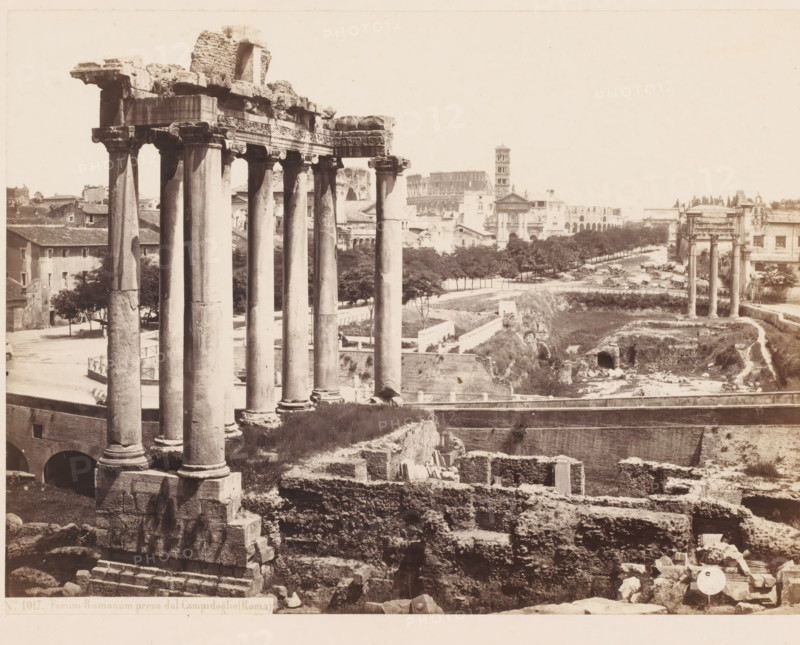 Forum Romanum preso dal Campidoglio (Roma), Ruins at the Roman Forum in Rome, Italy, photograph, inscription: 'Roma, Forum', address: 'SOMMER & BEHLES, ROMA E NAPOLI', number: '4', inscription: 'C. 85', number: 'No. 1017.', photographer: Sommer & Behles, Rome, 1867 - 1874, pasteboard, paper, albumen print, albumendruk, height 319 mm x width 380 mm, square, circus, ruin of a building, architecture, landscape with ruins, Forum Romanum, A wide sepia photograph shows a row of tall ancient stone columns with carved capitals in the foreground standing on large square bases, adjacent to fragmented stone ruins and terraced masonry; behind them a roadway lined with trees leads toward more classical ruins including another set of columns and a distant multi-level stone structure with arches and a tower, while surrounding the scene are low buildings, scattered stones, paths, and patches of grass and exposed earth.