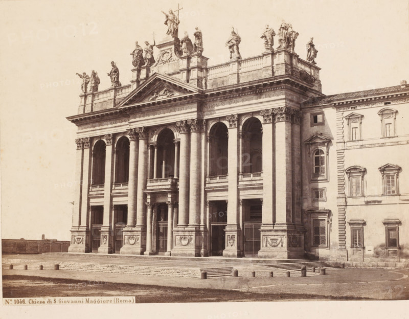 View of the Basilica of St. John Lateran in Rome, Italy, Chiesa di S. Giovanni Maggiore (Roma), photograph, number: '2.', inscription: 'Roma', address: 'SOMMER & BEHLES, ROMA E NAPOLI', number: 'No. 1046.', inscription: 'C. 85.', photographer: Sommer & Behles, Rome, 1867 - 1874, pasteboard, paper, albumen print, height 321 mm x width 382 mm, church (exterior), Sint-Jan van Lateranen, A sepia-toned photograph showing the façade of a large classical building with tall columns and arched openings, a wide flight of steps leading up to the entrance, and decorative relief panels at the column bases; a balustraded upper terrace topped by a row of statues standing on pedestals is visible along the roofline; adjacent to the main façade is a connected wing with rectangular windows and pilasters, and the foreground shows a paved open area with short cylindrical bollards and a textured surface.