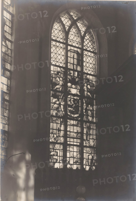 Painted window in the Castle Chapel of Egmond in Egmond aan den Hoef, photograph, annotation: 'stained glass window of Prince Frederik Hendrik, 1634 in the choir of the Dutch Reformed Church. Feb. 1907, Egmond a, d Hoef', annotation: 'Egmond a, d Hoef, stained glass window of Prince Frederik Hendrik 1634 in the choir of the Dutch Reformed Church, 1907', Castle Chapel (Egmond-aan-den-Hoef), photographer: AJM Mulder (possibly), 1907, Egmond aan den Hoef, Attributed to, paper, pasteboard, gelatin silver print, height 218 mm x width 149 mm, interior of church, church-window, e.g. rose-window, A tall arched stained glass window fills the center of the image, its upper sections showing diamond-patterned panes and curved mullions, while the lower panels feature decorative figurative and ornamental glasswork with human figures and heraldic motifs; light filters through the glass casting bright highlights against a dark interior wall, and a narrower stained glass window is partially visible alo...