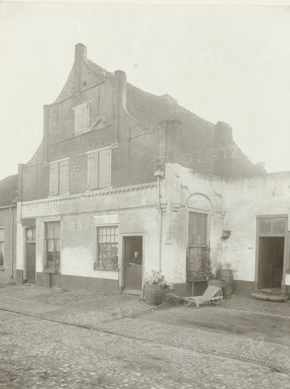 House in Amersfoort, Amersfoort 1905, photograph, title: 'Amersfoort 1905', name: 'H. Bickhoff Amsterdam', photographer: Henri Bickhoff, Amsterdam, 1905, pasteboard, paper, gelatin silver print, developing gelatin silver print, height 224 mm x width 172 mm, height 355 mm x width 298 mm, civic architecture, edifices, dwellings, Amersfoort, A two-story brick building with a stepped gable and tiled roof fills the scene, showing shuttered windows on the upper level and a mix of light and dark brickwork on the lower facade; a person is visible standing in a partially open doorway on the ground floor, next to a multi-pane window with objects on the sill and a small plant below; barrels and a wooden cart lie near the corner of the building on a cobblestone street, and a second doorway and arched window are visible along the right side, with some climbing vegetation on the wall.