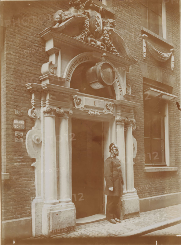Gate of the Spinhuis, now installed in the new police station in Spinhuissteeg in Amsterdam, photograph, annotation: 'Spinhuis', Amsterdam, 1910 - 1920, photographer, paper, pasteboard, daylight gelatin silver print, height 174 mm x width 126 mm, height 290 mm x width 226 mm, policeman, constable, civic architecture, edifices, dwellings, gate, entrance, Spinhuis, A sepia-toned photograph showing an ornate stone doorway set into a brick building, featuring carved columns with decorative capitals, curved scrollwork, and an arched pediment with sculptural ornament above. A round hanging sign projects from the arch, and a carved rectangular panel sits above the entrance. A man in a dark uniform and helmet stands at the doorway, facing sideways. To the right of the entrance is a window with a draped fabric decoration above it, and to the left a small mounted plaque on the brick wall. The street in front is paved with narrow bricks and a raised curb.