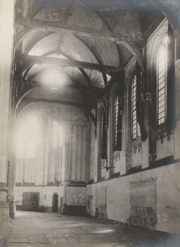 Interior of the Nieuwezijds Chapel in Amsterdam, photograph, annotation: 'Amsterdam, NZ Chapel, View into the side aisle, demolished Aug. 1908', photographer: Albert Adriaansz (possibly), Amsterdam, 1908, paper, pasteboard, gelatin silver print, developed gelatin silver print, height 216 mm x width 156 mm, height 325 mm x width 235 mm, interior of church, Nieuwezijds Chapel, A high vaulted interior with wooden arched beams crossing the ceiling and large tall arched windows along the right wall, light streaming through one window and creating bright flares. The walls show worn plaster and decorative stone corbels beneath the windows, with a ledge running along the right side. In the left and central area there are tall columns and slender arches leading toward a distant wall with more arched openings and a pair of wooden doors; one doorway is topped by a small rounded arch. Suspended metal chandeliers hang from the ceiling by long rods, and thin cables or wires descend along the walls. ...