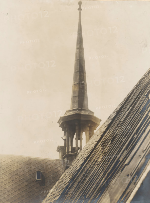 Tower of the Nieuwezijds Chapel in Amsterdam, photograph, annotation: 'Amsterdam, NZ Chapel, small tower, demolished 1908', photographer: Albert Adriaansz (possibly), Amsterdam, 1908, paper, pasteboard, gelatin silver print, developed gelatin silver print, height 212 mm x width 157 mm, height 323 mm x width 236 mm, parts of church exterior and annexes: tower - QQ - small church, chapel, Nieuwezijds Chapel, A tall, narrow spire rises from a stone base with an open, columned lantern beneath it, the spire showing segmented panels with weathered surfaces and a rounded finial at the top; adjacent sloped roofs with overlapping tiles occupy the foreground, one roof showing a small rectangular vent set into patterned shingles, and the image overall has a muted, aged tone with subtle stains and specks across the sky area.