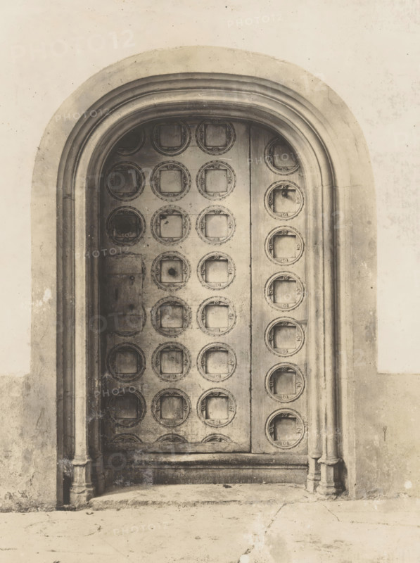 Portal in the Nieuwezijds Chapel in Amsterdam, photograph, annotation: 'Amsterdam, NZ Chapel, Door & door frame, preserved. Church demolished, Aug. 1908', photographer: attributed to Albert Adriaansz, Amsterdam, 1908, Attributed to, paper, pasteboard, gelatin silver print, height 190 mm x width 143 mm, height 0 mm x width 0 mm, door, parts of church interior (ENTRANCE), Nieuwezijds Chapel, A tall arched wooden door set into a stone frame with layered curved molding, the door surface covered in a grid of recessed circular panels each containing a square inset, the wood showing grain and worn textures with darker spots and scuffs, a rectangular metal plate and small round holes interrupting some panels, fluted stone columns at either side of the doorway rising from a stone threshold, a worn step and a textured ground in front, and a plain light-colored wall surrounding the arch with subtle stains and surface marks.