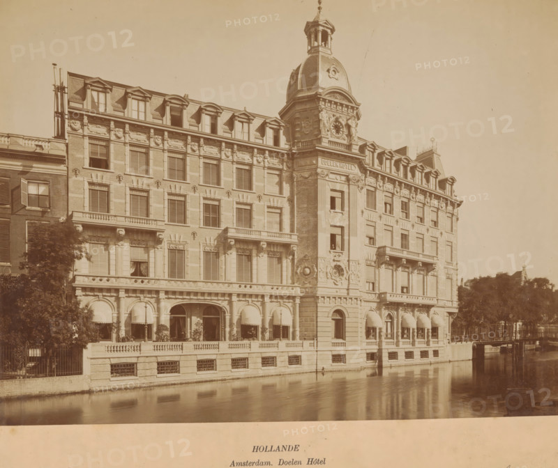 View of the Doelen Hotel in Amsterdam, photograph, inscription: 'Hollande, Amsterdam. Doelen Hôtel', annotation: 'Amsterdam', Amsterdam, 1883 - 1900, photographer, paper, pasteboard, albumen print, height 271 mm x width 280 mm, height 263 mm x width 332 mm, hotel, hostelry, inn, Doelenhotel, A wide sepia-toned photograph of a large ornate building along a canal, showing multiple stories with rows of windows, decorative stonework, and mansard-style dormer windows on the roof; the central section features a prominent tower with a domed roof, clock face, sculpted details, and a small open belfry at the top; the ground level has arched openings with striped awnings, columns, and a balustraded terrace with potted plants; the canal in the foreground reflects the building facade; trees and a fence appear at the sides, and a distant rooftop silhouette is visible on the right.