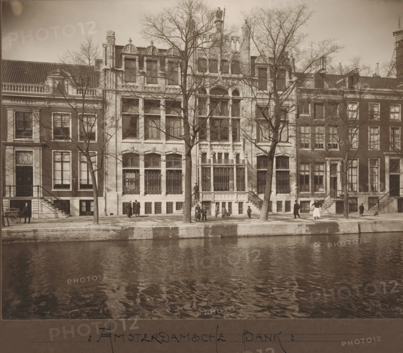 Facade of Herengracht 597 - 601 in Amsterdam, former Amsterdamsche Bank, photograph, annotation: 'Amsterdamsche Bank', annotation: 'Amsterdamsche Bank Heerengracht A'dam No 8, Architect Ed. Cuypers, Herengracht no. 8', inscription: 'Architect, Ed: Cuypers, Amsterdam', number: '1319, 164a', photographer: Fidel Carl Albert Greiner, Amsterdam, photographer: Albert Greiner, 1902 - 1930, Fidel Carl Albert Greiner, Albert Greiner, photographer, paper, pasteboard, daylight gelatin silver print, height 218 mm x width 276 mm, height 296 mm x width 420 mm, bank, street (city(-scape) with figures, staffage), façade (of house or building), Herengracht (Amsterdam), Amsterdamsche Bank, A row of tall buildings with ornate facades and large windows lines a tree-lined canal, with bare-branched trees standing in front of the central building and casting thin shadows on the stone sidewalk, decorative ironwork covering several lower windows and entrances, stone steps leading up to doorways, people standin...