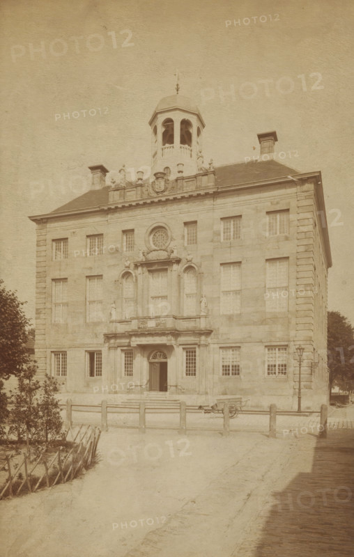 Enkhuizen Town Hall, photograph, cabinet photograph, annotation: 'VI, 1 c', inscription: 'Enkhuizen. Hotel de ville', Enkhuizen, 1870 - 1900, photographer, paper, pasteboard, albumen print, albumendruk, height 181 mm x width 115 mm, height 194 mm x width 123 mm, town hall, Stadhuis (Enkhuizen), A large stone building with a symmetrical façade rises from a paved approach, featuring evenly spaced rectangular windows and decorative stonework around a central entrance with steps and a recessed doorway framed by columns and an arched transom. Above the entrance is an ornate balcony area with sculpted figures and a circular window set into the wall, and a small dome-like cupola sits atop the roof with open arches. Chimney-like structures project from the roof on either side, and a low wooden fence runs across the foreground beside a dirt path. A lamppost stands near the building's right side, a cart is positioned near the entrance, and trees and trimmed hedging appear at the left and right e...