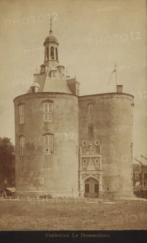View of the Drommedaris in Enkhuizen, photograph, cabinet photograph, annotation: 'VI, 1a', inscription: 'Enkhuizen Le Drommédaire', Enkhuizen, 1870 - 1900, photographer, paper, pasteboard, albumen print, albumendruk, height 179 mm x width 115 mm, height 194 mm x width 123 mm, harbour, Drommedaris (Enkhuizen), A large round brick building with two cylindrical towers connected by a curved wall occupies the image, showing multiple tall rectangular windows and a decorative arched entrance with sculpted stonework; a steep slate roof rises to a cupola with arched openings and a bell visible inside, topped by a finial and weather vane; a flagpole stands on the roof near a small chimney; a grassy foreground with short posts and a fence runs across the base of the building, and partial neighboring structures and trees are visible at the sides.