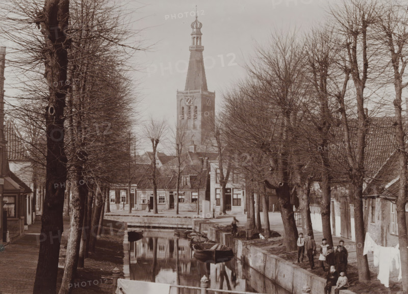 View of Medemblik with the tower of the Reformed Church, photograph, inscription: 'B. Reekers, Photographer, Enkhuizen', annotation: 'View of the tower of the Reformed Church in Medemblik, Photo B. Reekers', annotation: 'N:6, 13 1, 2 wide', photographer: Barend Reekers, Medemblik, 1890 - 1920, paper, pasteboard, daylight collodion silver print, height 126 mm x width 173 mm, height 141 mm x width 186 mm, canals, waters (in city), church (exterior), street (city(-scape) with figures, staffage), A tree-lined street runs beside a narrow canal with small rowboats moored along the stone edge, bare branches of the trees forming a lattice against the sky; buildings with pitched roofs and multiple windows face the water, and a tall church tower with a steep spire and an ornate top rises above the rooftops; people stand and sit along the canal bank and near the buildings, and a line of laundry hangs between trees, all reflected in the still surface of the canal.