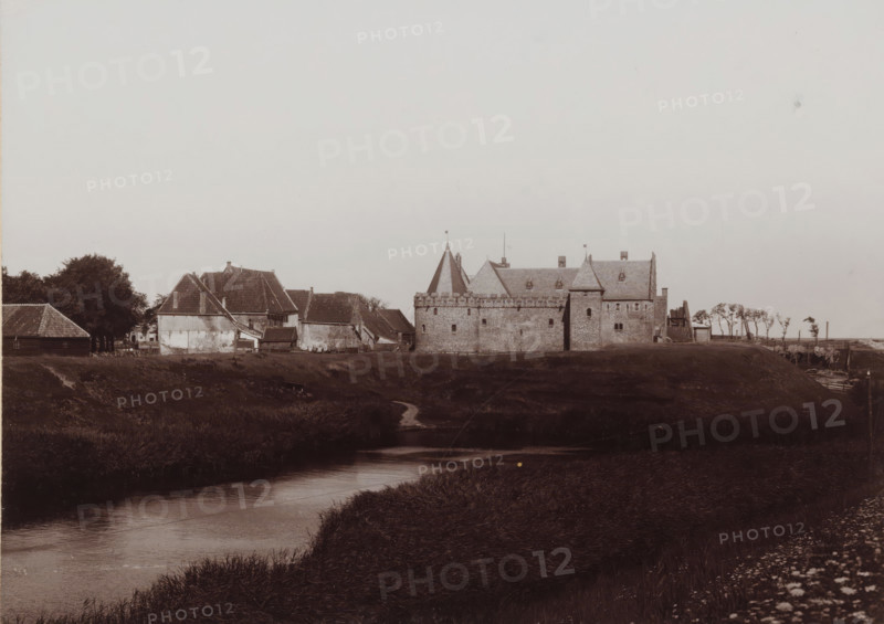 View of the former Koggenhuis in Medemblik, photograph, annotation: 'Medemblik, Former Koggenhuis in Medemblik', inscription: 'B. Reekers, Photographer, Enkhuizen', photographer: Barend Reekers, Medemblik, 1890 - 1915, paper, pasteboard, daylight collodion silver print, height 121 mm x width 167 mm, height 173 mm x width 126 mm, urban housing, Radboud Castle, A wide view of a low-lying landscape with a meandering waterway in the foreground and grassy banks leading toward a row of buildings on the far bank, including masonry structures with battlemented walls and steeply pitched roofs, a conical tower with a small flag, and adjacent cottages with tiled roofs and weathered exterior walls, clusters of trees near the buildings, a narrow dirt path running from the bank down to the water, and a strip of open sky above.