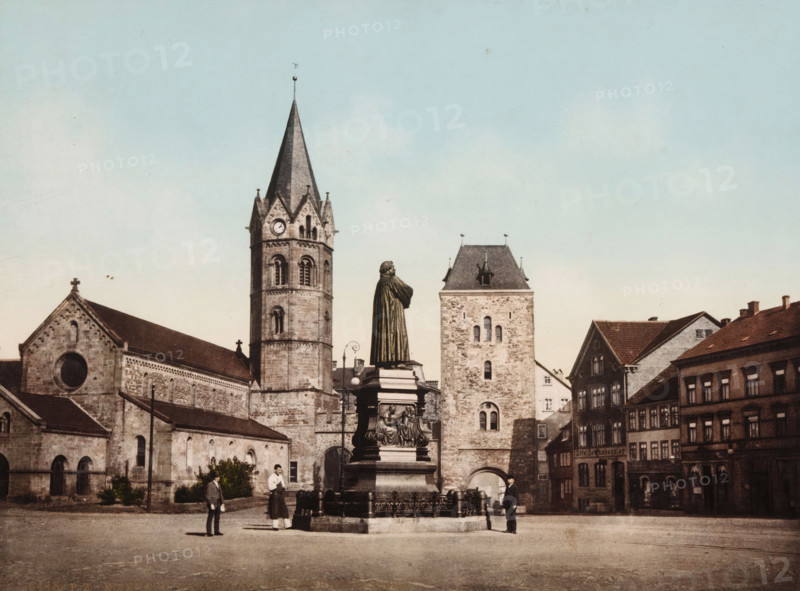 View of the Karlsplatz in Eisenach with the statue of Martin Luther, Eisenach, Karlsplatz mit Lutherdenkmal, photomechanical print, number: '6448.', cliché maker: Photochrom Zürich, Zürich (city), 1889 - c. 1920, photographer, Thüringen, pasteboard, paper, photochrom, height 188 mm x width 248 mm, Person, Martin Luther, monument, statue, square, circus, Karlsplatz, A stone plaza scene with a tall statue of a robed figure on an ornate pedestal surrounded by a low decorative fence, a large church building with a tall pointed bell tower and visible clock face, an adjacent square stone tower with small arched windows, and rows of multi-story buildings with pitched roofs; a few people stand near the statue and along the plaza, and the sky above is pale blue.