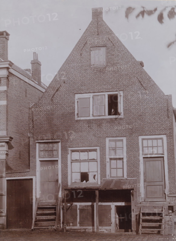 Façade of Buitenveer 141 in Weesp, photograph, annotation: 'Buitenveer 141, demolished Nov. 1911, Weesp', Weesp, photographer: AJM Mulder (possibly), 1910 - 1911-11, Attributed to, paper, pasteboard, daylight collodion silver print, height 210 mm x width 154 mm, urban housing, Buitenveer, A brick gabled building with a steep triangular roof and a small boarded opening near the top stands centered in the image, showing patterned brickwork and a chimney at the apex; a rectangular window with one open pane is visible below the midpoint, and beneath that are several framed windows and doors with peeling paint and broken glass, some set above wooden steps and small platforms supported by posts; adjoining brick structures with chimneys and decorative trim appear at the left and right edges, and tree leaves are visible at the top right against a pale sky.
