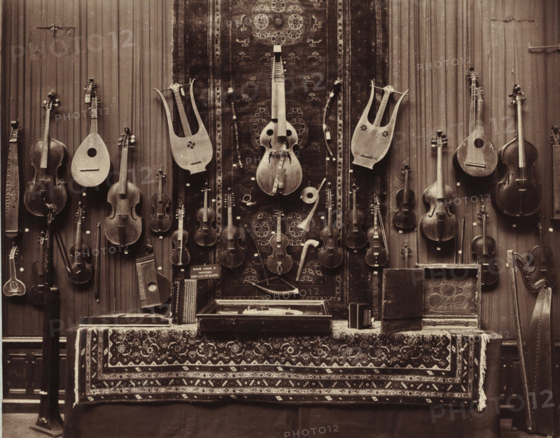 Musical instruments from the Daniël François Scheurleer collection, exhibited in Pulchri Studio in 1893, photomechanical print, inscription: 'A. vd GRIENT, PHOT. - THE HAGUE.', photographer: Adrianus van der Grient, Pulchri Studio, 1893, The Hague, pasteboard, paper, Woodburytype, Woodburytypie, height 221 mm x width 280 mm, Person, Daniël François Lunsingh Scheurleer, exhibition, art, string instruments (plucked), musical instrument, A wall covered with a patterned hanging and vertical wooden panels displays a variety of stringed instruments suspended at different heights, including several violins, cellos, a double bass, lyres, lutes, and other plucked and bowed instruments with visible tuning pegs and carved bodies; a large carved harp stands to the right edge. Below the instruments a table draped with an ornate patterned textile holds open cases, a small box, and other accessories, while a long rug with repeating geometric motifs hangs centrally behind the instruments, showing worn...