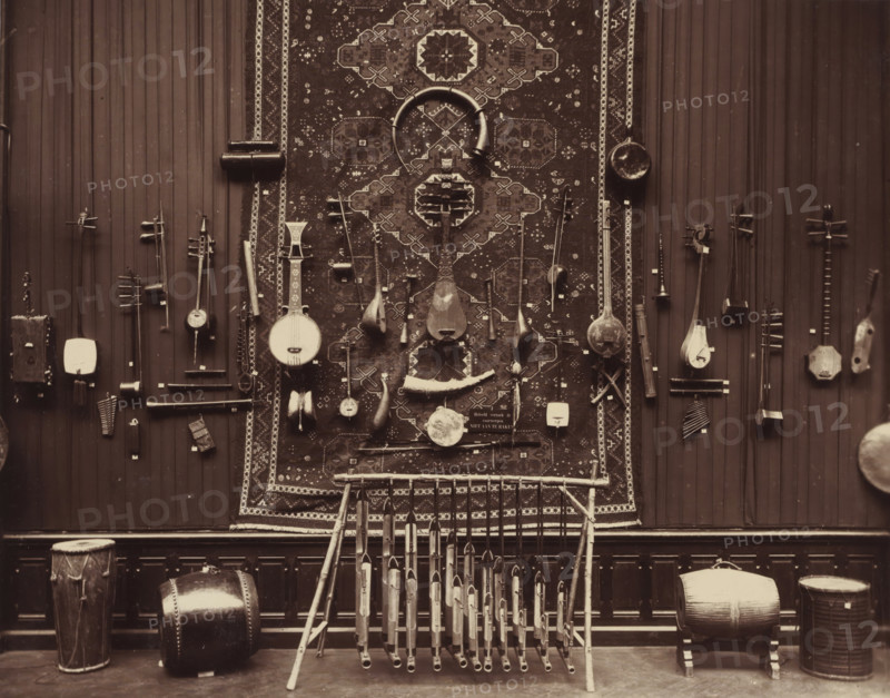 Musical instruments from the Daniël François Scheurleer collection, exhibited in Pulchri Studio in 1893, photomechanical print, inscription: 'A. vd GRIENT, PHOT. - THE HAGUE.', photographer: Adrianus van der Grient, Pulchri Studio, 1893, The Hague, pasteboard, paper, Woodburytype, Woodburytypie, height 220 mm x width 280 mm, Person, Daniël François Lunsingh Scheurleer, musical instruments, group of musical instruments, exhibition, art, musical instrument, A large patterned rug hangs centrally on a vertical paneled wall, with a variety of stringed and percussion instruments mounted around and on it; several long-necked lutes and rounded-bodied instruments hang from the rug and wall, some with carved necks and pegboxes, and smaller hand-held instruments with curved shapes are interspersed; a pair of crescent-shaped objects and a circular drumhead are visible on the rug; numerous slender pipes arranged in a wooden frame stand in front of the rug, their open ends facing outward; drums of d...