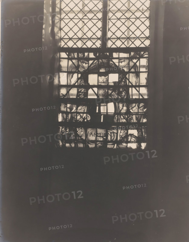 Painted window in the Castle Chapel Egmond in Egmond aan den Hoef, photograph, annotation: 'Egmond a, d Hoef, stained glass window of Purmerend 1634 in the south facade of the Dutch Reformed Church, Feb 1907', annotation: 'stained glass window of Purmerend in the south facade of the Dutch Reformed Church, Feb. 1907, Egmond a, d Hoef', Castle Chapel (Egmond-aan-den-Hoef), 1907, photographer, Egmond aan den Hoef, Attributed to, paper, pasteboard, gelatin silver print, height 205 mm x width 163 mm, interior of church, church window, e.g. rose window, A tall window with diamond-patterned leaded panes fills the upper center of the image, showing a grid of lighter glass panels framed by darker mullions; behind the panes there are decorative painted or stained sections with figurative shapes and ornamental motifs visible through the lower portion of the window, and darker vertical wall surfaces flank the window on both sides, while the lower two-thirds of the image is dominated by a large dar...