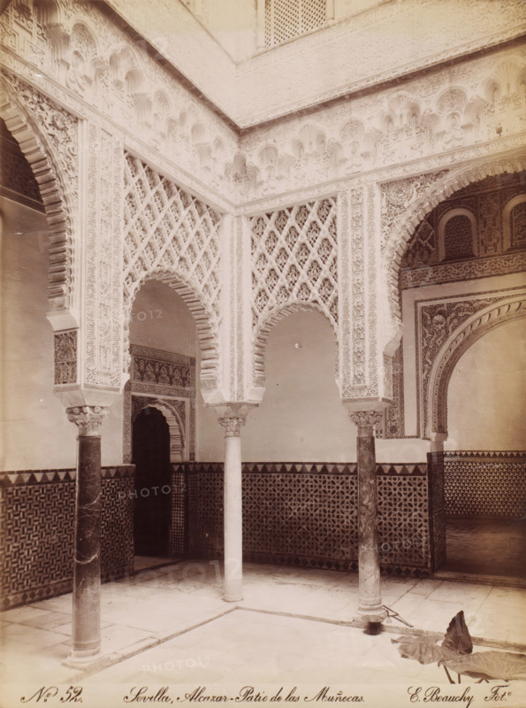 Courtyard of the Alcazar in Seville, No. 52, photograph, Seville, Alcazar - Patio de las Muñecas., photographer: Emilio Beauchy, c. 1867 - c. 1880, pasteboard, paper, albumen print, height 386 cm x width 290 cm, palace, Andalusia, Royal Palace of Seville, A stone courtyard corner with ornate carved arches and patterned wall tiles, showing slender columns with decorative capitals supporting horseshoe-shaped arches, intricately carved plaster latticework and geometric reliefs above and between the arches, a tiled dado with repeating star and polygon patterns, a smooth paved floor with recessed channels, a dark recessed doorway beneath one arch, and a large fallen leaf lying on the floor near one column.