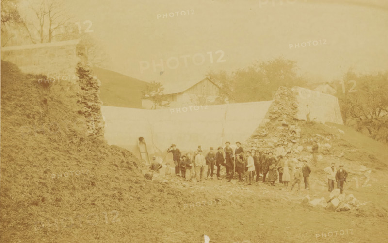 Workers at a construction site, photograph, photographer: Edouard Boulland, Vevey, c. 1880 - c. 1900, pasteboard, paper, albumen print, albumendruk, height 332 mm x width 402 mm, building-site, anonymous historical persons depicted in a group, in a group-portrait, A faded sepia photograph showing a group of people standing and posing on rough, rocky ground in front of a low wall and a pile of stones, with a sloped grassy embankment on the left and trees and simple gabled buildings in the background; the figures include men, women, and children wearing period clothing and hats, some holding tools or leaning on walking sticks, arranged in loose clusters across the central foreground, while a couple of individuals stand apart near the right edge and another person stands on the stone pile.