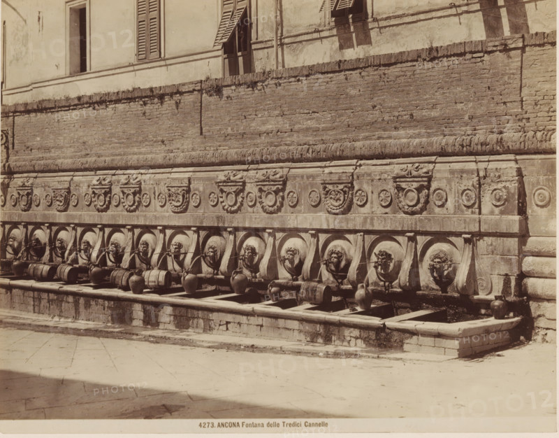ANCONA Fontana delle Tredici Cannelle, Fountain of Calamo in Ancona, photograph, number: '4273.', Ancona, c. 1875 - c. 1900, photographer, Marche, paper, pasteboard, albumen print, albumendruk, height 189 mm x width 239 mm, ornamental fountain, A stone wall with decorative carved reliefs runs across the image, above a row of circular basins each set beneath a sculpted lion head spout; clay jars and wooden barrels sit in the basins and on the ledge below, while water trickles from the spouts into the basins; above the wall there is a building facade with shuttered windows and an open doorway, and the wall shows weathering and patches of vegetation; the foreground is a paved stone plaza with light and shadow across its surface.